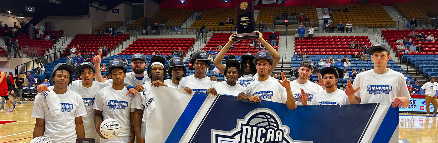 JALC men's basketball team posing with awards and NJCAA Division 1 Men's Basketball Championship banner