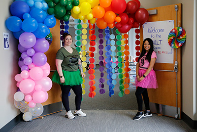 Two female nursing students dressed in fun outfits, next to a door decorated with colorful balloons at the Dr. Barb Patchett Eggstravaganza.