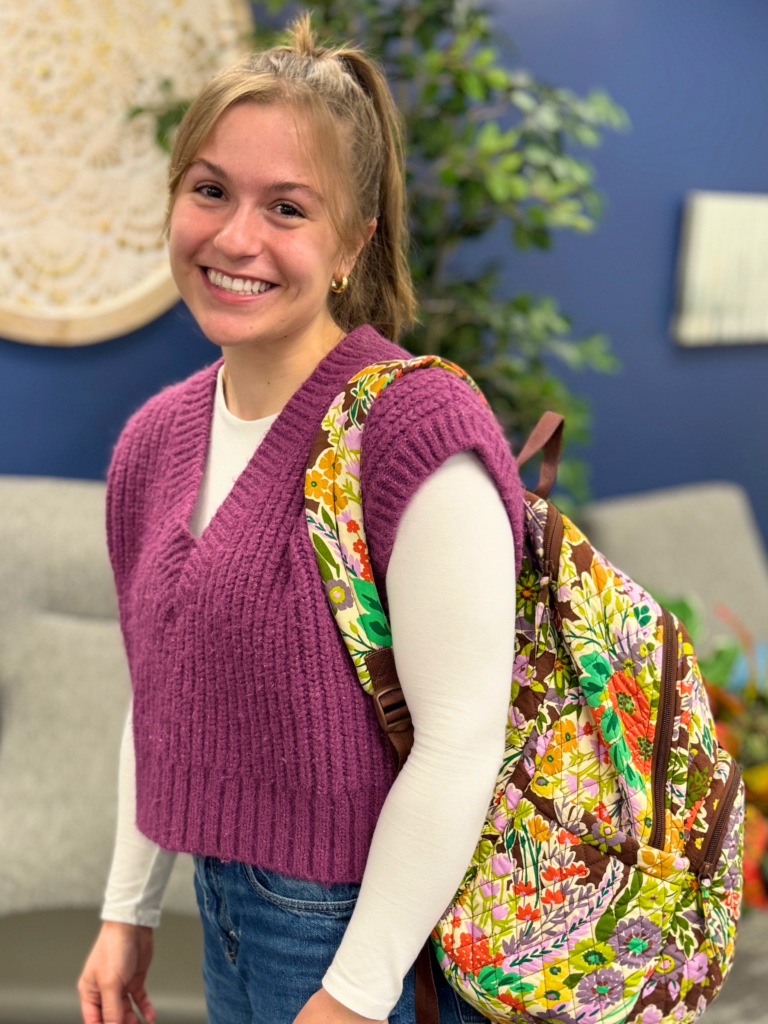 Jacie Nunley standing with her colorful backpack over her should smiling for a picture wearing a pink sweater and white shirt with a ponytail in front of a blue decorated wall