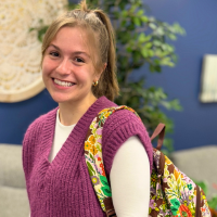 Jacie Nunley standing with her colorful backpack over her should smiling for a picture wearing a pink sweater and white shirt with a ponytail in front of a blue decorated wall
