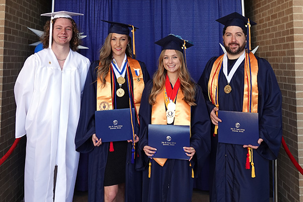 Two male and two female students posing for a picture wearing blue graduation regalia with cords, medals, and sashes with their diplomas.