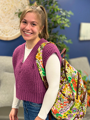 Jacie Nunley standing with her colorful backpack over her should smiling for a picture wearing a pink sweater and white shirt with a ponytail in front of a blue decorated wall.