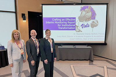 Dr. Stephanie Chaney Hartford, Emily Monti, and Joseph Dethrow stand next to screen with their presentation.