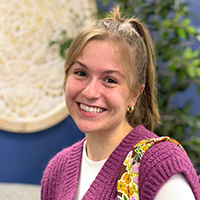 Jacie Nunley standing with her colorful backpack over her shoulder smiling for a picture wearing a pink sweater and white shirt with a ponytail in front of a blue decorated wall.
