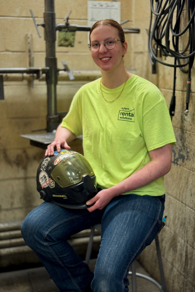Caroline Farthing is seating in a welding stall holding her welding helmet posing for a picture wearing a lime green Xylem shirt