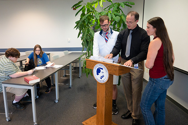 JALC Instructor of Political Science and Sociology Matt Lees works with two students at a podium while two other students work at a table in a classroom.